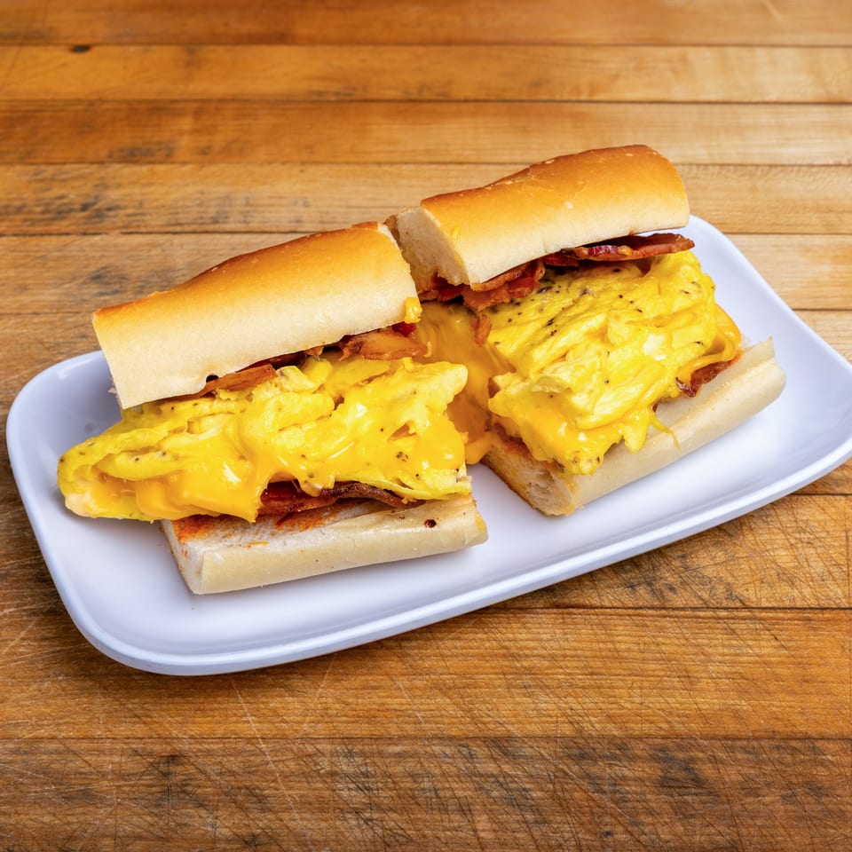 Two bacon, egg, and melted cheese sandwiches on toasted rolls, served on a white plate atop a wooden table.