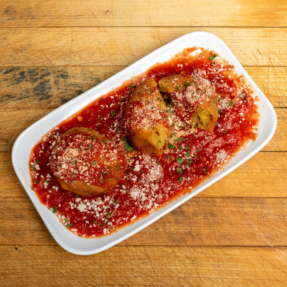 Breaded stuffed peppers served in tomato sauce, topped with crumbled cheese and chopped parsley on a white platter.