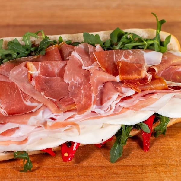 A large prosciutto and mozzarella sandwich with arugula and roasted red peppers on a wooden board.
