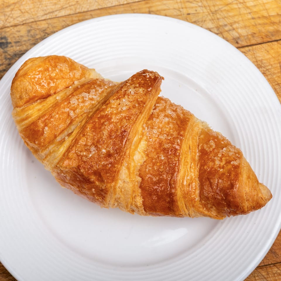 Golden, sugar-topped croissant on a white plate atop a well-used wooden cutting board.