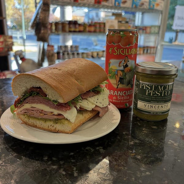 A large Italian sandwich with meats, cheese, and pesto, next to a blood orange soda and pistachio pesto jar.