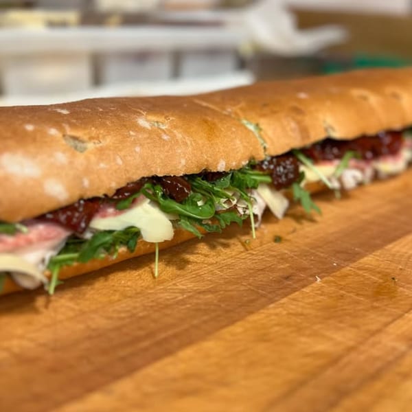 A long baguette sandwich filled with arugula, cheese, salami, and sun-dried tomatoes on a wooden board.