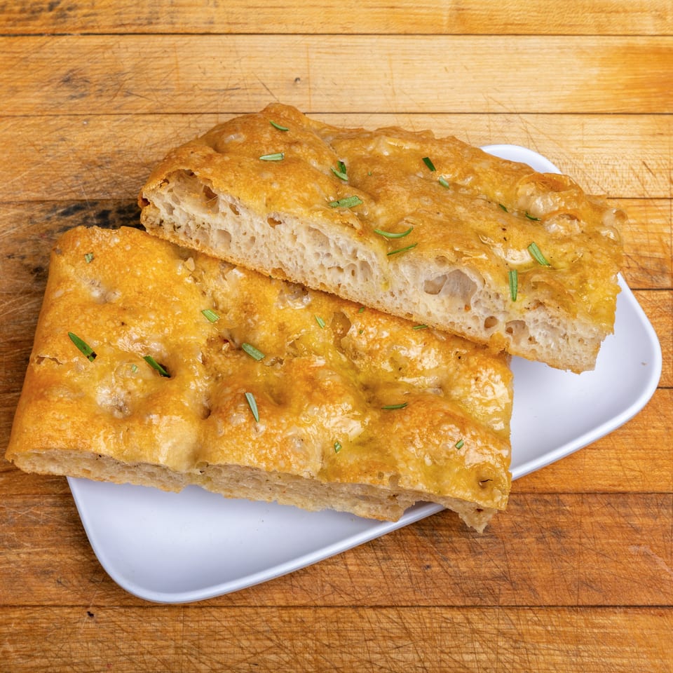 Golden-brown focaccia bread topped with herbs, sliced and stacked on a white plate over a wooden cutting board.