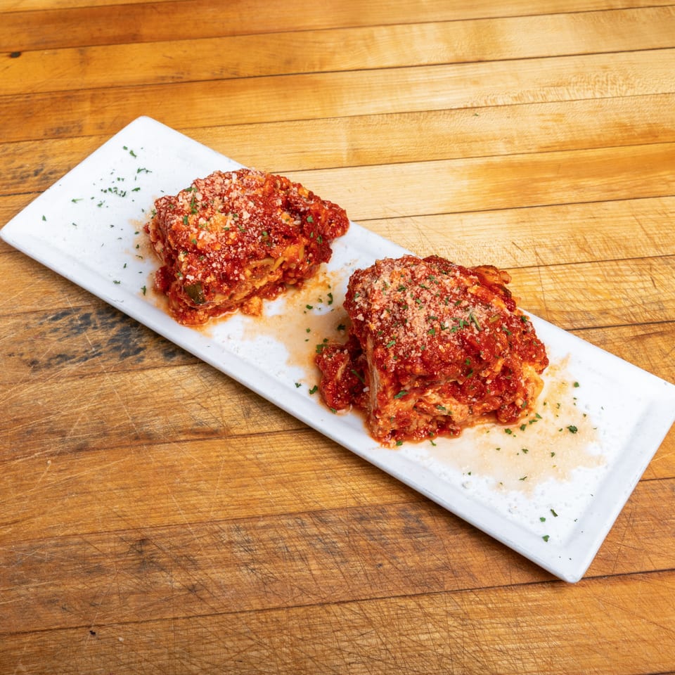 Two slices of lasagna topped with tomato sauce, grated cheese, and parsley on a white platter over a wooden table.
