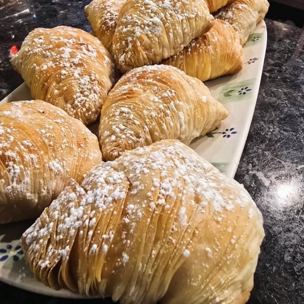A platter of golden-brown sfogliatelle pastries dusted with powdered sugar on a dark countertop.